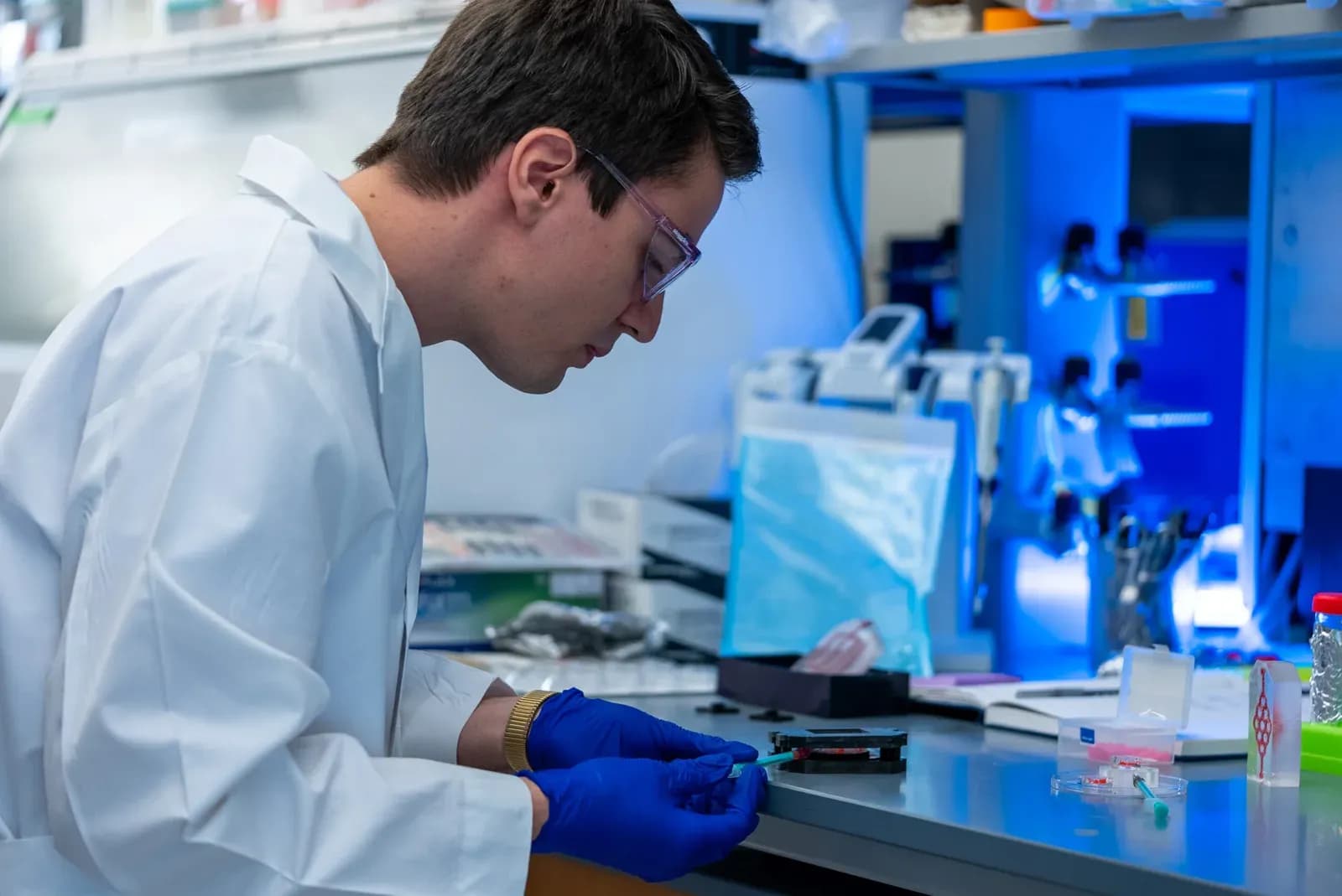 A researcher in a white lab coat, safety glasses and blue gloves works at a laboratory bench, handling a small sample holder with a pipette beside blue-lit equipment.
