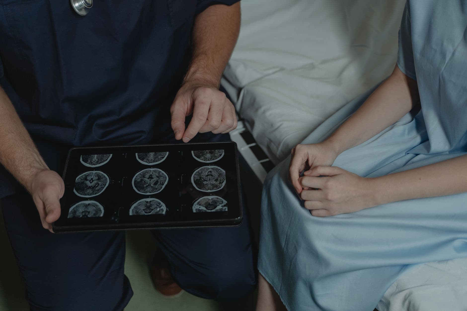 A clinician in scrubs shows brain scan images on a tablet to a seated patient in a hospital gown.