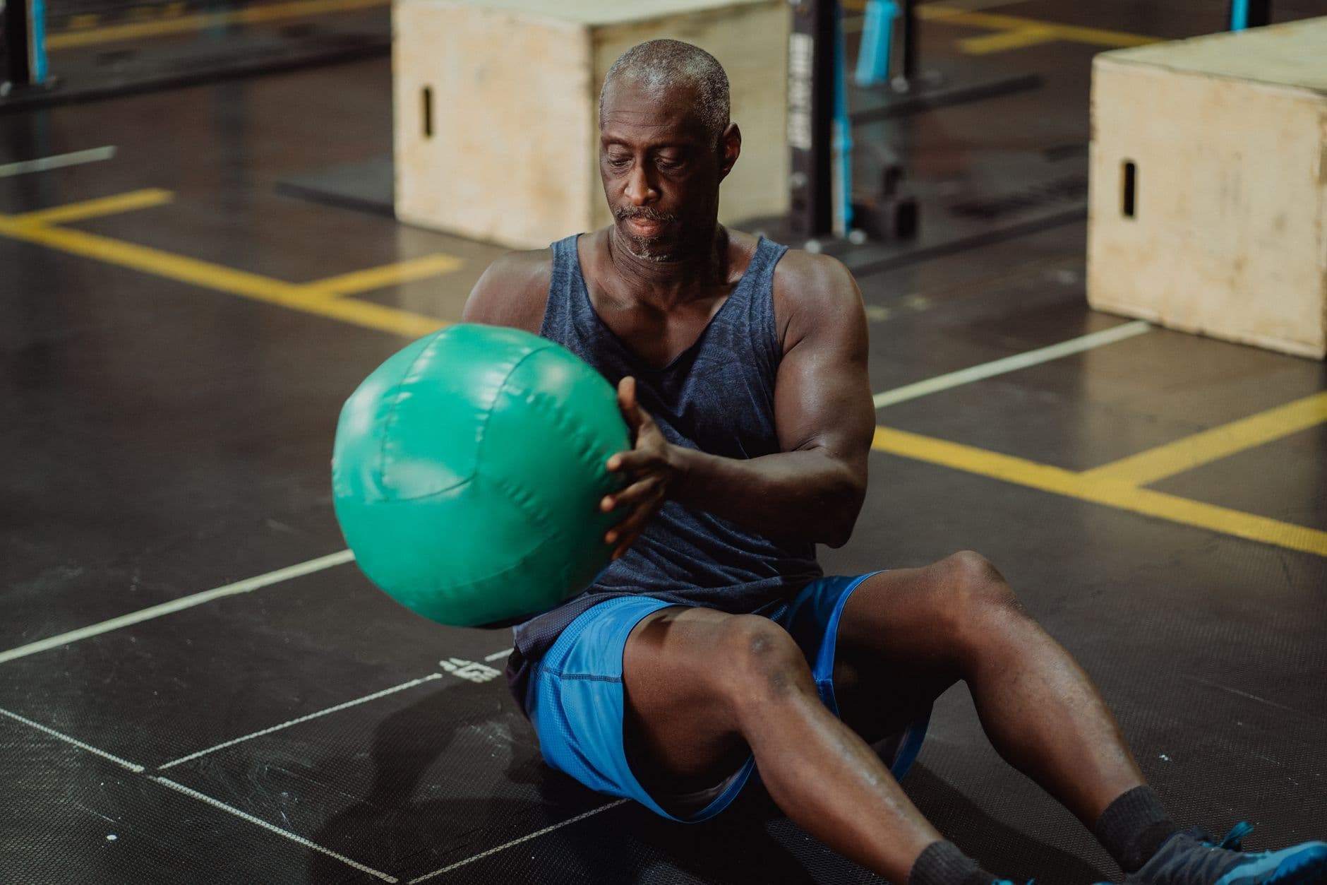 A mature man seated on a gym floor doing a controlled medicine ball exercise