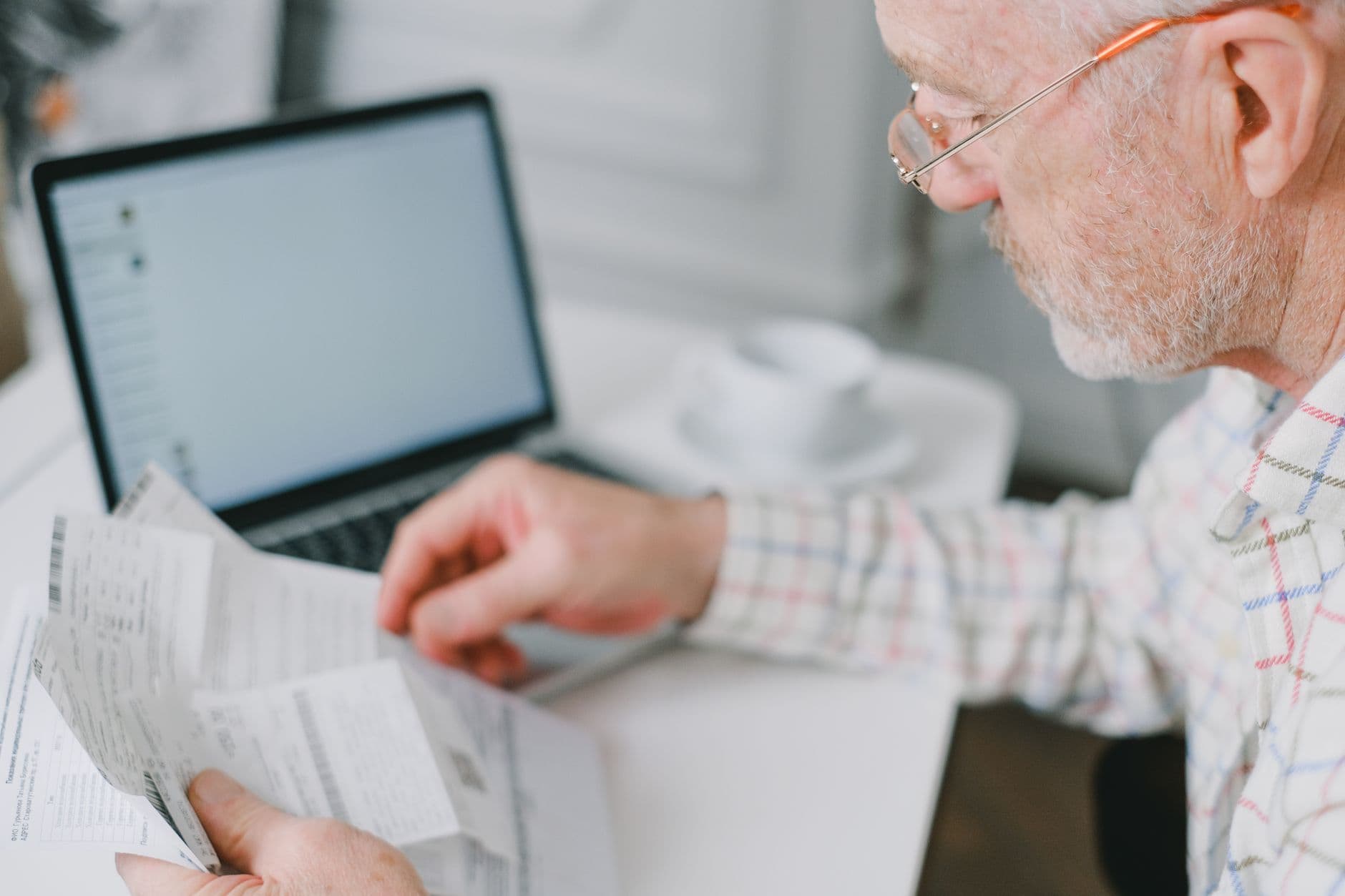 Elderly man wearing eyeglasses reading documents next to a laptop at home.