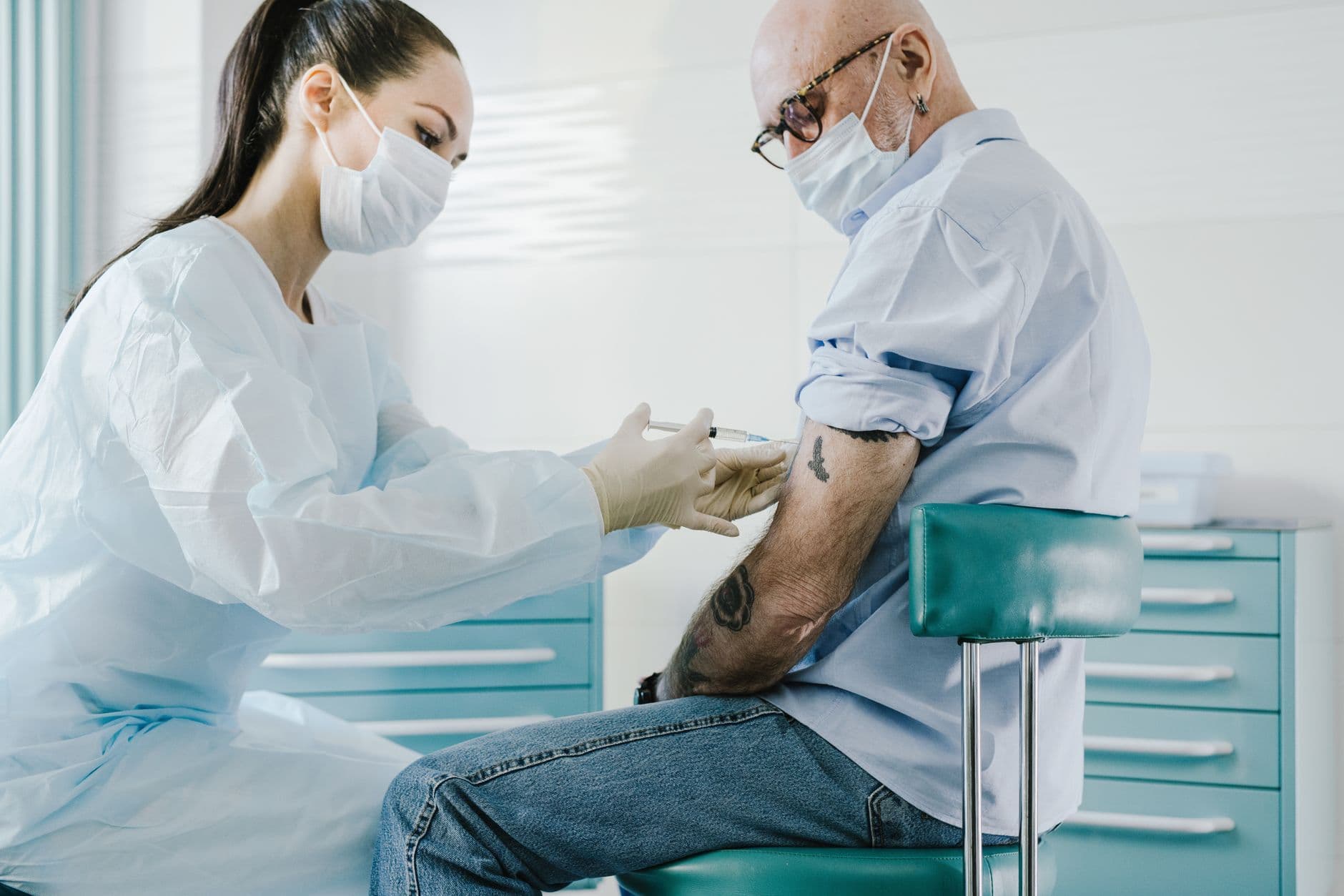 A nurse wearing a mask and gloves gives a vaccine injection into the upper arm of an older man seated in a clinic.