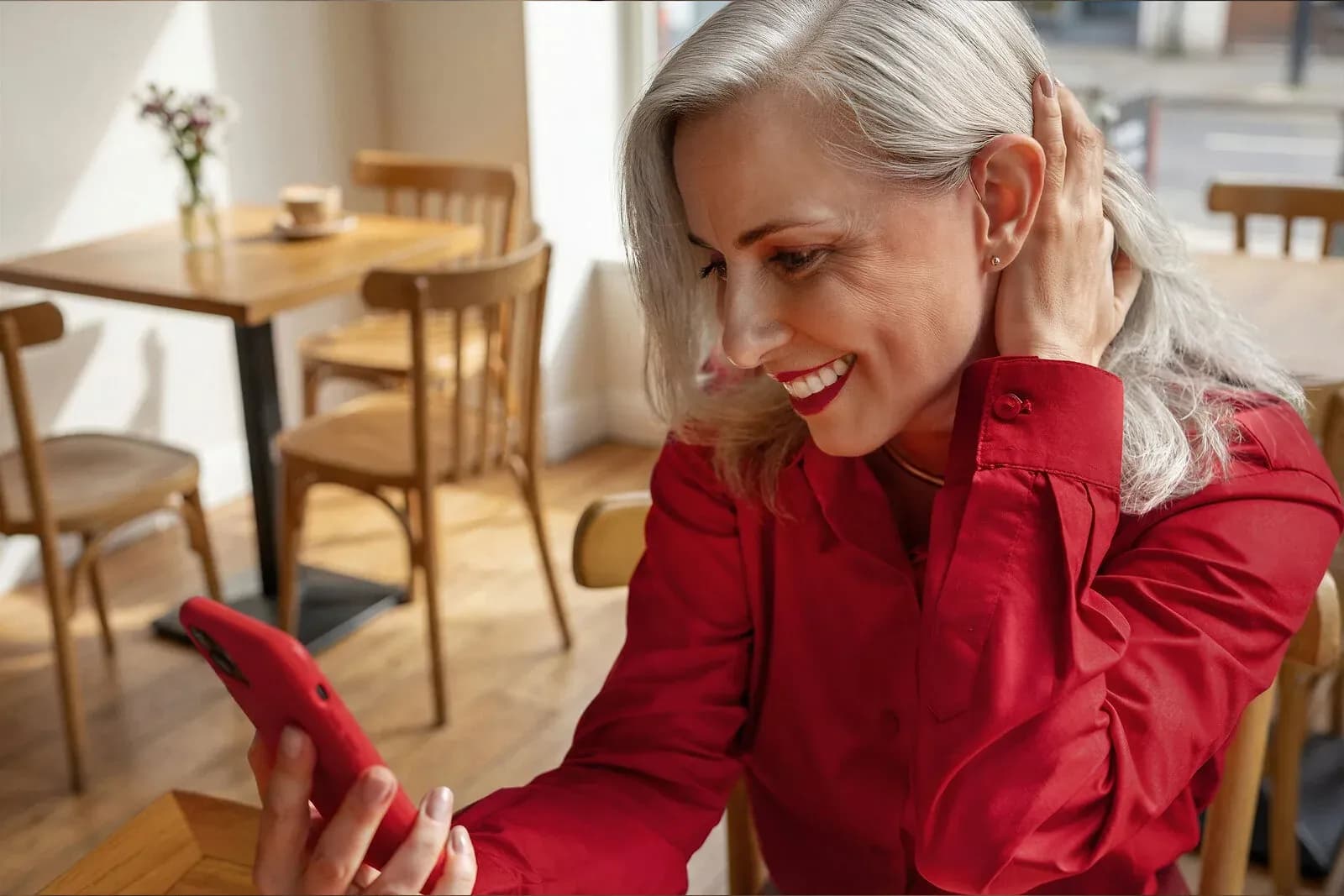 A silver-haired woman smiles at her phone in a cafe, one hand near her ear