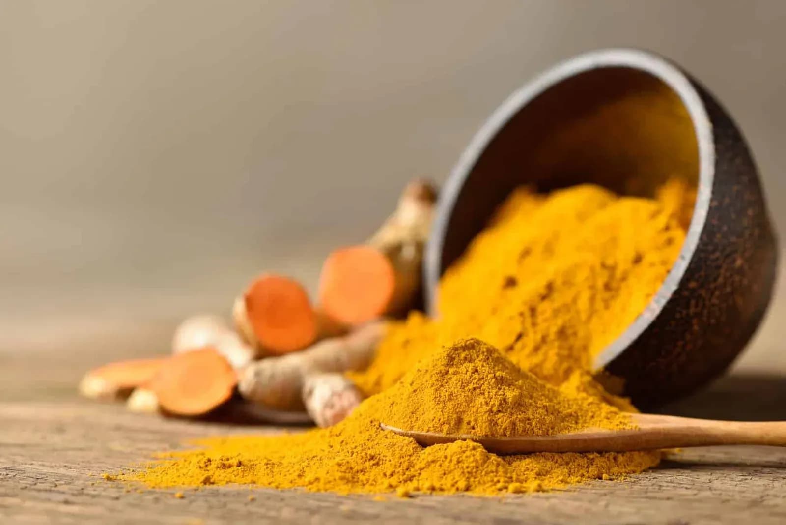Ground turmeric spilling from a dark bowl beside cut turmeric root on a wooden board.